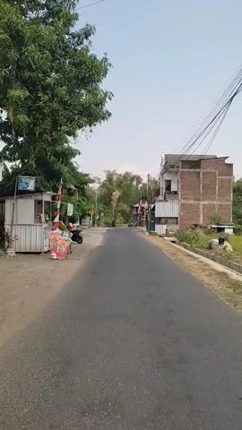 A person is walking down a paved road in a rural area. On the left side of the road, there is a small stall with some products displayed. Further down the road, there are trees and some buildings. On the right side of the road, there are houses and a large brick building. The sky is clear and blue. There are some wires running overhead. The person appears to be walking away from the camera, towards a gate-like structure in the distance. The overall impression is one of a typical village scene.