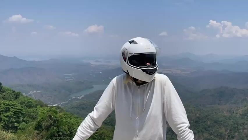 A person wearing a white motorcycle helmet and a white hoodie stands with their arms slightly outstretched in front of a scenic mountain view. The helmet has a clear visor that is flipped up, revealing a dark inner visor. A small logo that appears to read "Royal" is visible on the helmet. The background features layers of green, tree-covered mountains under a blue sky with scattered white clouds. A body of water is visible in the distance, nestled between the mountains.