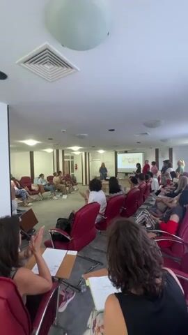 The video shows a presentation in a room with about twenty people seated in red chairs facing a projector screen. The projector screen displays a colorful graphic with what appear to be portraits of people. A woman is standing at a podium on the left side of the room, gesturing and speaking. Several other people are standing near the screen, some of whom appear to be interacting with the presenter. The room has white walls, fluorescent lighting, and wooden accents. There is a whiteboard on the left wall with writing on it. The audience members are diverse in age and gender, and many are looking towards the front of the room. Some audience members have notebooks and pens.