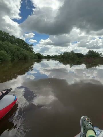 A static, eye-level shot from a watercraft shows a river under a partially cloudy sky. The water surface reflects the sky, creating a mirror effect. The river banks are lined with green trees and bushes. A person's feet are visible at the bottom of the frame, one wearing black sandals and the other wearing green Crocs with a charm. A red paddle rests near the feet.