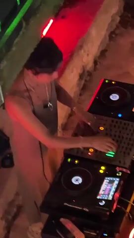 A person wearing a black beanie and a tan sleeveless dress is standing behind a DJ setup, consisting of two turntables and a mixer. The person is holding a bottle of water in their left hand and interacting with the mixer with their right hand. Red and green lights are visible in the background, casting a colorful glow on the scene. The environment appears to be outdoors, possibly a party or event setting, with a stone wall in the background.