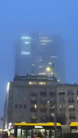 A tall building, partially obscured by fog, is the main focus of the video. The building has multiple visible signs, including "WELL FITNESS", "biznes zone", "ATLAS TOWER", and "TWÓJ START UP". The building is illuminated with lights from within, giving it a hazy glow through the fog. In the foreground, there is a smaller, older building with visible windows and balconies. A street is present in the lower part of the frame, with a yellow bus driving by. The environment appears urban and the weather condition is foggy.