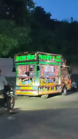 The video shows a food truck at dusk. The truck is brightly lit with neon lights and decorated with colorful images of food items. There are people in the background and a scooter parked nearby. The truck appears to be stationary.