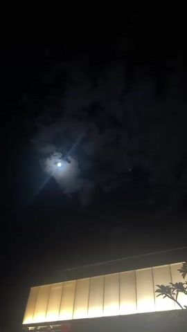 This vertical video clip captures a nighttime scene with a bright full moon visible through scattered clouds. The moon is the dominant subject in the sky, with a lens flare emanating from it. The lower portion of the frame shows the illuminated facade of a building, suggesting an urban or semi-urban setting. The building's windows are not visible, but the entire front appears to be lit with warm, yellow light, creating a glowing effect. A silhouetted tree branch is visible on the right side of the frame, adding to the nighttime ambiance. The overall mood is serene and somewhat mysterious, highlighted by the contrast between the bright moon and the dark sky. The time of day is clearly night, and the presence of clouds indicates a possibly humid or temperate climate. No people are visible in the frame. There is no visible text in the video.