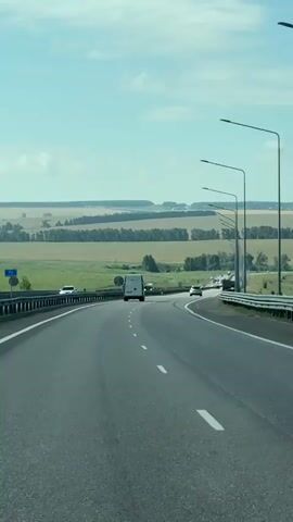 The video shows a view from a vehicle traveling on a highway. The road is paved and has white dashed lines indicating lanes. There are streetlights lining the right side of the road. On the left side of the road, there is a guardrail and grassy embankment. In the distance, there are rolling hills and fields of crops, with some trees and scattered buildings. Several vehicles are visible on the road, including cars, a van, and trucks. The sky is blue with scattered white clouds. The camera perspective suggests the video was recorded from the driver's or passenger's seat of a moving vehicle.