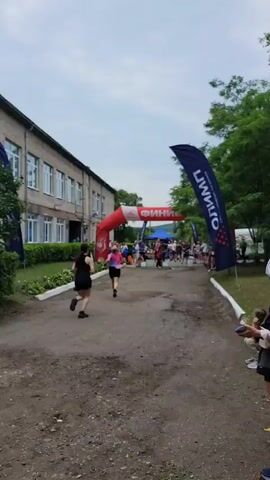 A group of runners are crossing a finish line under a red inflatable archway. The archway has the word "ФИНИШ" (FINISH in Russian) written on it in white letters. In the foreground, a woman in a black tank top and black shorts is running towards the finish line. Behind her, a woman in a pink and black shirt and black shorts is also running. Spectators are visible on both sides of the finish line. A blue tent is set up behind the archway. The scene appears to be outdoors, with trees and a building visible in the background. The sky is overcast.
