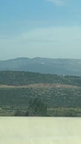 The video shows a car driving on a road, with a landscape view in the background. The landscape consists of green, hilly terrain with a city visible in the distance under a hazy blue sky. A white "hybrid" car pulls up to the side of the road. It has an Israeli flag attached to the back of the vehicle. A lamppost is visible next to the car.