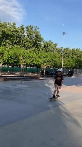 A young boy, wearing a white helmet, a dark short-sleeved shirt, light-colored shorts, and black sneakers, is skateboarding in an outdoor skatepark on a clear, sunny day. He starts on a flat concrete surface, with lush green trees and a bright blue sky with scattered clouds in the background. Distant cars and other skatepark structures, including ramps, are visible.

The boy rolls forward on his skateboard towards an orange metal rail. He attempts to ride over the rail, but as the front of his board clears it, his back foot and the rear of the skateboard hit the rail. He loses balance and steps off the skateboard with his left foot, landing on the concrete ground beside the rail. The skateboard tumbles to the other side of the rail. The boy then stands next to the rail, looking at his fallen skateboard. The word "LIBIANO" is spray-painted on a concrete wall behind the rail.