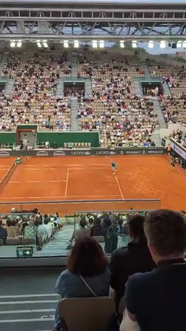 The video depicts a tennis match being played on a clay court in a large stadium. The stadium is packed with spectators filling the stands. The court is orange and bordered by advertisements. The camera is positioned at a high angle from the stands, providing a wide view of the scene. One player is visible on the court, moving to return a shot.  A glass barrier separates the camera from the court, with spectators seated directly behind it. The stadium has a visible roof structure and is brightly lit.