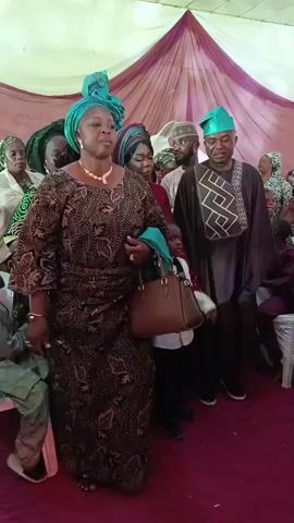 A group of people, primarily adults and a few children, are standing and moving slightly at what appears to be an event. The central figure is a middle-aged woman in an intricately patterned brown dress with a turquoise headwrap and purse. She is wearing jewelry and has a serious expression. She is positioned slightly ahead of the group and appears to be the focus. The others are wearing traditional African clothing in various colors. A small boy is holding her hand, dressed in a white shirt and bow tie. The background is a tent-like structure with maroon and white draping, and there are white chairs scattered around. The flooring visible is red. Music is playing in the background.