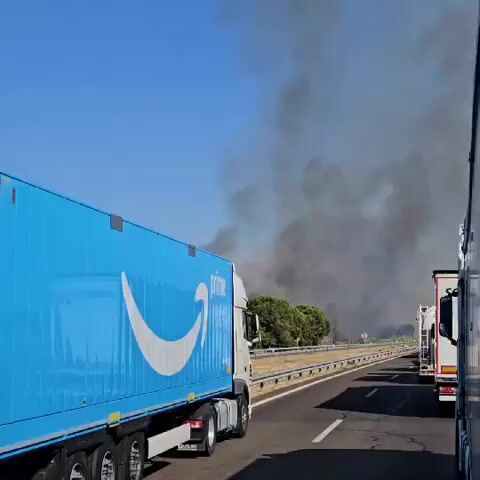 The video shows a view from a moving vehicle on a highway. In the foreground, a large blue truck with an Amazon Prime logo is visible. The truck is in the left lane. To the right of the Amazon truck, other trucks are also visible in the same lane. Beyond the highway, there is a stretch of dry grass and trees. A large plume of thick, dark smoke is rising from behind the trees, indicating a fire. The sky is clear and blue. The overall scene suggests traffic on a highway near a location where a fire is burning.