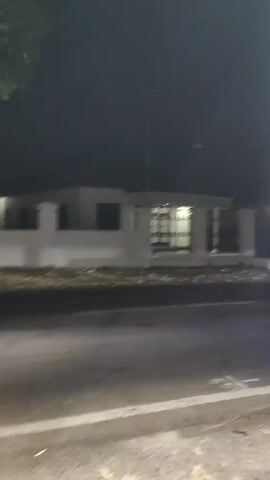 The video shows a nighttime scene from a moving vehicle. The camera pans across a street with trees and buildings on the side. One building appears to be a light-colored, single-story structure with a dark roof and windows. Another structure is a yellow and red container. A car with its headlights on drives past the camera. The road is paved and appears to be a street in a town or city. The lighting is dim, characteristic of nighttime.