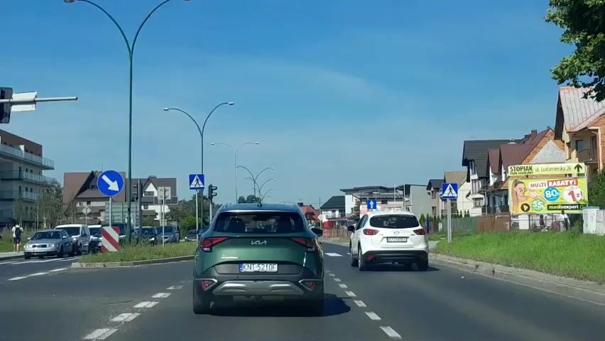 A dashboard camera view shows a dark green Kia Sportage driving on a two-lane road on a sunny day. A white Kia Sportage is ahead of the camera vehicle, also driving in the same lane. On the left side of the road, there are cars parked along the side and apartment buildings. On the right side of the road, there are houses and a billboard advertising a telecommunications company. There are pedestrian crossings and traffic lights visible in the video. The camera vehicle is driving on a paved road with white lane markings.