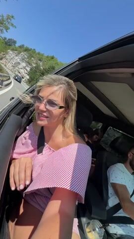 The video shows a woman in the passenger seat of a car, leaning out the window with her arm raised. She is wearing a pink and white striped off-the-shoulder top and sunglasses. She appears to be singing or talking, with her mouth open and head tilted back. The car is driving on a winding road with a mountainous landscape in the background. Another person is visible in the back seat of the car. The sun is shining and the sky is blue.