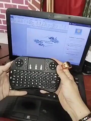 The video shows a person holding a black wireless mini keyboard with a touchpad and several directional buttons. The person is using the keyboard to interact with a laptop that is open. The laptop screen displays a document with a decorative border of blue leaves and text that appears to be in a formal font. The laptop is on a dark surface, possibly a desk, with a brick-patterned wall in the background. The person is wearing a gold ring on their right ring finger. The video is shot from a close-up perspective, focusing on the hands and the keyboard.