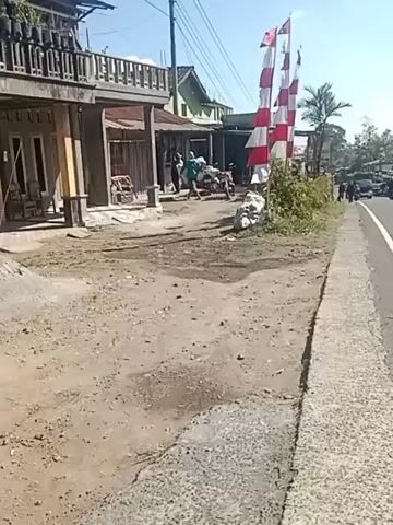 The video shows a dirt road in front of a building with a balcony. There are several people present, some carrying items. Red and white flags are visible, possibly indicating a celebration or national holiday. A motorcycle is parked near the building. The road appears to be unpaved and somewhat uneven. In the background, there are other buildings and vehicles, suggesting a village or small town setting. The sky is clear and the weather appears to be sunny. The overall impression is that of an everyday scene in a rural or semi-rural area.