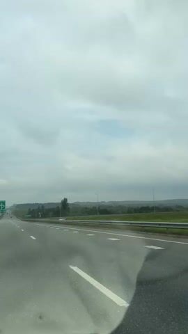 The video shows a view from a vehicle traveling on a highway. The sky is overcast with clouds. On the left side of the road, there are green highway signs and other signage. The highway itself has multiple lanes, marked with white lines. To the right of the highway, there is a metal guardrail and then a grassy area with trees and foliage. In the distance, there are more trees and rolling hills. The overall environment appears to be rural or suburban. The camera is static, indicating it is mounted within the moving vehicle. There are no people or other vehicles clearly visible in the frame.