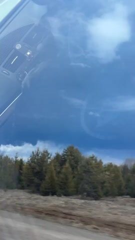 The video, filmed from inside a moving vehicle in Yugo-Kamskiy, Russia, shows a vast landscape under a partly cloudy, somewhat dark sky. Reflections of the car's dashboard, including air vents and controls, are visible in the top portion of the frame.

The vehicle is traveling along a road with bare fields and scattered coniferous trees on either side. In the distance, a massive, dark smoke plume rises high into the sky, suggesting a significant fire or industrial event. Power lines and utility poles are also visible across the landscape.

Road signs are seen along the route: one indicating a turn or merge (an arrow pointing up and right), and another displaying a speed limit of "50" kilometers per hour, with a smaller sign below it that includes "100m". The overall impression is of a drive through a rural area where a large-scale event is unfolding in the distance.