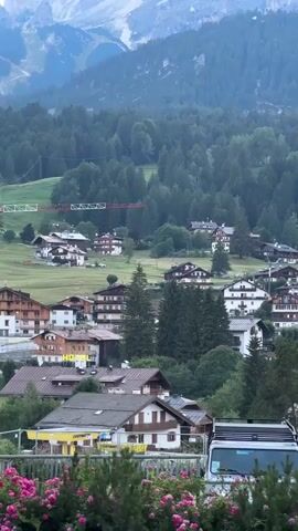 The video opens with a panoramic view of a mountain village nestled amidst a lush green forest, with towering, rugged mountains in the background. The camera pans upward, revealing the majestic peaks, their slopes partially covered in snow. The village itself is characterized by traditional alpine architecture, with numerous chalets featuring wooden balconies and dark roofs. A few ski lifts are visible on the mountainside, hinting at the area's recreational activities. The foreground is adorned with vibrant pink flowers and a glimpse of a white vehicle. The scene suggests a tranquil mountain setting, possibly during the summer or early autumn.