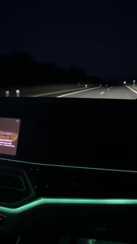 The video shows the interior of a car at night. The dashboard has green ambient lighting. The car is on a highway, and trees are visible on the side of the road. The car's display shows what appears to be album art and song information for a track titled "Russian DMX". The camera is stationary, pointing forward from inside the car.