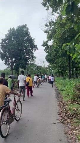 The video shows a group of people walking and riding bicycles down a paved road that is lined with trees and vegetation on both sides. The sky is overcast. In the foreground, a person on a bicycle is partially visible on the left. Further down the road, a group of individuals are walking and cycling away from the camera. One person is wearing a white t-shirt and grey pants. Another person is wearing a yellow bag over their shoulder and red pants. Several other people are walking and riding bicycles, some of them appearing to be children. The road appears to be a rural or suburban area.