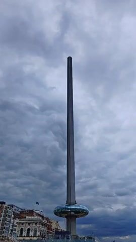 The video shows a tall, slender tower with a pod-like structure at its base. The tower is silhouetted against a cloudy sky. The architecture of buildings can be seen in the distance. The scene appears to be outdoors during the daytime. There are sounds of people talking in the background.
