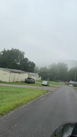The video is shot from the perspective of someone in a vehicle driving down a wet asphalt road. The sky is overcast and foggy, obscuring the distant hills and trees. To the left of the road, there is a building with a light-colored exterior and a dark roof, with a gray pickup truck parked in front of it. Further down the road, a silver sedan is parked. The road appears to be in a rural or suburban area, with houses and trees visible on either side. The overall atmosphere is calm and subdued due to the weather conditions.