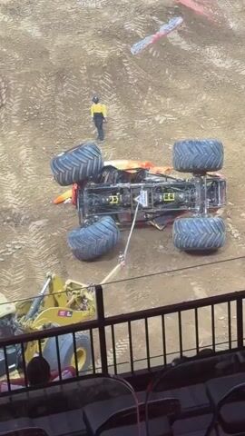A monster truck, "El Toro Loco," is being righted by a front-end loader. The monster truck is upside down, with its wheels in the air. A person in a yellow shirt and black pants stands near the monster truck. Another person is partially visible in the lower left corner of the frame, seemingly operating the front-end loader. The environment appears to be a dirt arena.