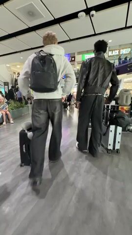 Two individuals walk through an airport terminal. The person on the left has blonde hair and wears a white hoodie, dark jeans, and a black backpack. They are pulling a black suitcase with a neck pillow draped over its handle. The person on the right has dark hair and wears a black leather jacket, dark pants, and white sneakers. They are pulling a silver suitcase. In the foreground, a suitcase with rainbow-colored stripes is visible. The background shows other travelers, escalators, signage, and airport amenities. The flooring is a polished gray.
