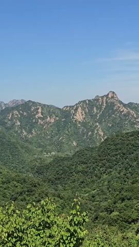 The video pans across a mountainous landscape covered in lush green trees under a clear blue sky. As the view moves, a section of the Great Wall of China comes into view, with several people walking along its parapets. The wall is made of stone and brick and is situated on a ridge overlooking the surrounding forest and distant mountains.
