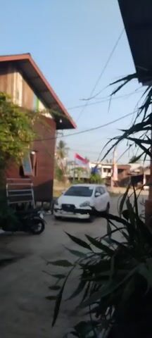 The video shows a white car parked in a dirt driveway. To the left of the car is a wooden house with a red roof. A motorcycle is parked next to the house. In the background, there are other houses and trees. An Indonesian flag is flying on a pole behind the car. The sun is shining and the sky is blue. The video is shaky and out of focus.
