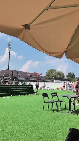 The video shows an outdoor scene on a sunny day. In the foreground, part of a large, light brown umbrella is visible. Beneath the umbrella, on a bright green lawn, are two black metal chairs and a small matching table. To the right of the table, a person in dark swimwear is standing. Further back, on the lawn, several people are scattered around. Some are lying on lounge chairs, others are standing or walking. A man in dark shorts is walking away from the camera towards a building. To the left, there are some green steps leading up. Behind the lawn and people, a long, low building with a white facade and a red sign that reads "Weekend" is visible. A tall cell tower stands to the left of the building. The sky is blue with scattered white clouds. The overall impression is a relaxed outdoor recreational area, possibly a beach club or pool resort.