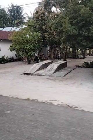 The video shows a street scene in a village. A white house with a brown roof is on the left. In front of the house, there is a sandy yard with some trees and bushes. A road runs along the right side of the frame. The video pans across the scene, showing more houses and shops. There are also some motorcycles and bicycles visible. The sky is overcast. The video appears to be taken from a moving vehicle.