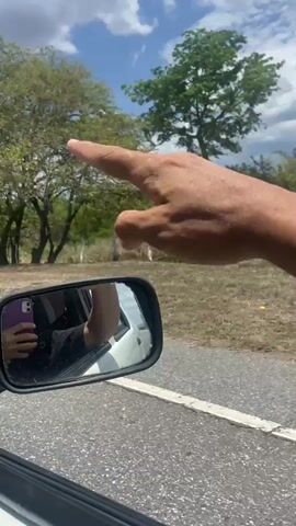The video offers a close-up view from inside a moving vehicle, looking out the right-hand passenger window. A person's right hand, adorned with a ring on the ring finger, is extended out of the car window. The car's side mirror occupies a significant portion of the foreground, reflecting the person's left hand holding a mobile phone with a purple case, along with a partial view of their upper body dressed in dark clothing and the white exterior of the car.

Throughout the short clip, the right hand makes several pointing and gesturing movements with its index finger, appearing to playfully interact with the external environment or its reflection in the mirror. This could be a creative act of "framing" distant objects or drawing shapes in the air.

The setting is a rural or semi-rural landscape in Orupe, Venezuela. A paved road is visible, flanked by green trees and dry, grassy ground. In the background, under a bright, partly cloudy blue sky, distant structures and utility poles can be seen. The clear daylight indicates a sunny day. The overall activity conveys a sense of playful engagement with the passing scenery during a drive. No discernible text is visible in the video.