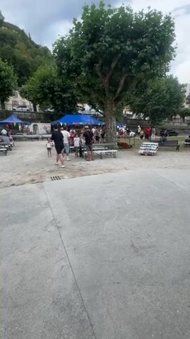 The video shows an outdoor gathering with several people present. In the foreground, there is a concrete area with some benches and tables set up. Beyond that, there is a dirt or gravel area where more people are mingling. Several large trees with green foliage are scattered throughout the scene, and a hill with trees and buildings can be seen in the background. The sky is overcast with clouds. There are also some blue tents and white canopies set up, suggesting a market or event. One person is riding a small red bicycle across the concrete area. In the middle ground, a group of people are standing together, and some are sitting at tables. Further back, more people are visible under the tents. Near the end of the video, a close-up shot reveals a speaker on a stand and some electrical cords near a green structure that appears to be a covered pavilion or bandstand.