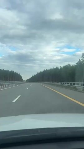 The video shows a view from the front of a car driving on a highway. The highway is flanked by a dense forest of tall pine trees on both sides, stretching as far as the eye can see. The road has two lanes in each direction, separated by a concrete barrier and guardrails. White lane markings are visible, along with a solid yellow line marking the right edge of the driving lane. The sky is cloudy with patches of blue peeking through, suggesting a somewhat overcast but potentially clearing day. The time of day appears to be daytime, given the brightness and the visibility of the road and surroundings. The overall mood is one of travel and perhaps a sense of solitude or immersion in nature. The location is identified as Demikhovo, Russia. There is no visible text in the video.
