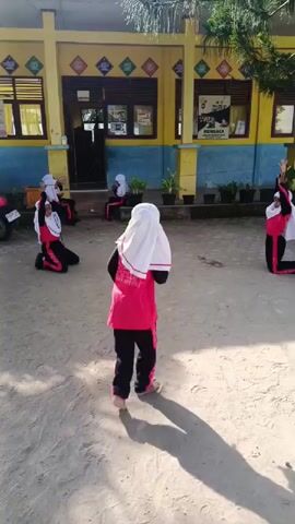 The video captures an outdoor scene at what appears to be a school in Simpangjaya, Indonesia. Five young girls are visible, dressed in matching attire: pink/red long-sleeved tops, black pants with pink accents, and white hijabs. They are engaged in a synchronized dance or exercise routine on a sandy, dirt ground.

The girls demonstrate various poses and movements; some are kneeling with their arms raised, while others are standing and stepping, their arms gently moving. Their actions suggest a coordinated performance or practice session.

In the background, a brightly painted building with yellow upper walls and blue lower walls stands prominently. It features multiple windows, some adorned with decorative patterns. A visible sign on the building reads "MEMBACA," which is Indonesian for "READ," reinforcing the school setting. Potted plants are neatly arranged along the base of the building.

The scene is bathed in bright sunlight, with clear shadows cast on the ground, indicating a mid-morning or early afternoon time. The overall atmosphere is one of focused activity and perhaps joyful engagement in a physical education or extracurricular event. A partial view of a motorcycle or bicycle is seen on the far left of the frame.