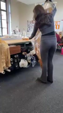 A woman in a grey workout suit is in what appears to be a sewing or design studio. She is taking pieces of a light gold colored fabric from a clear plastic bag and placing them on a table next to a mannequin that has red lines marked on it. There are rolls of various colored fabrics underneath the table. The room has white walls and a few large windows that let in natural light. There are more mannequins and sewing stations in the background.