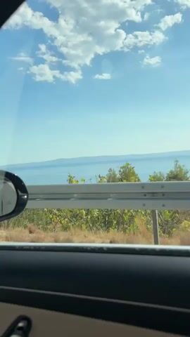 The video shows a view from inside a car, looking out the passenger side window. The car is traveling on a road, indicated by the movement of the scenery. The landscape outside consists of trees, dry grass, a metal guardrail, and in the distance, a body of water, likely the sea, under a bright blue sky with scattered white clouds. The sun appears to be shining, creating a bright and clear day. The interior of the car is visible at the bottom of the frame, showing part of the car door and seat. The view is dynamic, implying the car is in motion, with trees and vegetation passing by. There are no people or animals visible in the frame.