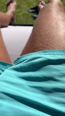 A first-person perspective video shows a person relaxing on a lounge chair outdoors. The person is wearing turquoise swim trunks and their legs are prominently featured. Their bare feet are visible at the top of the frame, with a pair of black flip-flops resting on the grass behind them. The background consists of a well-maintained green lawn, lush trees, and a brick wall. The sky above is a bright blue with fluffy white clouds, suggesting a sunny day.