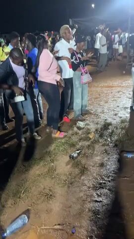 The image depicts an outdoor nighttime gathering, likely a party or concert, in Osogbo, Nigeria. A diverse group of young adults is present, illuminated by bright artificial lights that cast shadows. The ground is muddy and wet, suggesting recent rainfall, and is littered with discarded items including an empty dark glass bottle, red plastic cups, and white paper or wrappers.

In the foreground, several individuals are prominent. A woman in a light pink long-sleeved top and black leggings is dancing energetically, with her arms raised and a joyful expression. Beside her, a man with light-colored hair, wearing a white polo shirt, is also dancing with his arms up and a wide smile. To the right, another woman in a black crop top, patterned wide-leg pants, and a black beanie is holding a pink crossbody bag while looking at her phone. On the far left, a woman in a denim jacket, purple top, and light jeans, carrying a white crossbody bag, sways gently. Faint white text, possibly "MN," is visible on the dark pants of a person standing to the left.

In the background, numerous other attendees stand, watch, and mingle, with white tents or canopies visible further back under the dark sky. The overall atmosphere appears festive and energetic, with many participants expressing enjoyment and engagement.