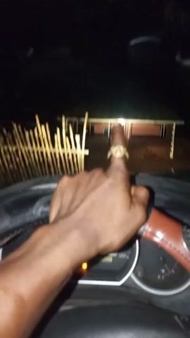 The image shows a view from inside a car at night, looking out through the windshield. A person's right hand is visible on the steering wheel, which has a two-tone brown and black cover with some textured elements. The car's dashboard is in the foreground, displaying a speedometer, tachometer, and several illuminated indicator lights, including engine, battery, and brake warnings. The odometer reads "217203".

In the background, a single-story house is visible, illuminated by a light at its front. To the left of the house, there is a rustic wooden fence. The ground in front of the house appears to be unpaved. The scene is dark, indicating it is nighttime, with the car's headlights casting light forward. The car appears to be stationary. The setting suggests a rural or semi-rural environment, consistent with the provided context of Iyara, Nigeria.