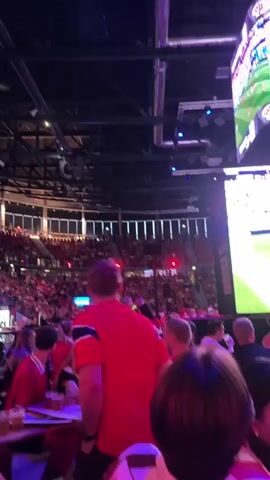 This video shows a large group of people gathered indoors, likely in a stadium or arena, watching a live broadcast of a soccer match on multiple large screens. The main subjects are the spectators, who are predominantly wearing red shirts, suggesting they are fans of a particular team. Many are seated at tables, some with drinks and food, indicating a social viewing experience. The atmosphere appears lively and engaged, with people looking towards the screens, which display a soccer field with players in action.

The scene is set in a dimly lit indoor venue with a high ceiling, characteristic of a sports arena. The screens are bright and prominently displayed, showing the game from different angles. In the foreground, people are visible in focus, with the background showing more spectators in tiered seating, all bathed in the glow of the screens and ambient lighting.

The activity is clearly the collective viewing of a soccer game. The time of day is not explicitly shown, but the indoor setting and artificial lighting suggest it could be evening or a daytime event held indoors. There are no explicit weather cues. Location cues are limited, but the presence of the text "BESTILL MATER" and "DJURSTRAK" on one of the screens, coupled with the prompt specifying Oslo, Norway, strongly suggests the location is in Norway. The emotions conveyed are excitement and shared interest, typical of a sporting event's audience.

Visible text includes "BESTILL MATER" (Order Food) and "DJURSTRAK" on one of the large screens, which are likely advertisements or informational displays related to the venue or event. The overall impression is of a lively and communal sports viewing event in a modern indoor stadium in Oslo.