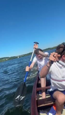 Three people are in a canoe on a sunny day. The sky is clear and blue. The water is choppy and dark. They are paddling near a red buoy. The man in the middle is laughing as he paddles, and another man paddles in the front. They are wearing light-colored, casual clothing. There is land visible in the distance.