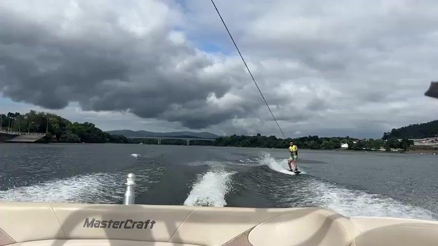 A wakeboarder is being pulled by a boat on a lake. The wakeboarder is wearing a yellow life vest and green shorts. The boat is a MasterCraft. The water is choppy and there is a large wake behind the boat. The sky is cloudy. There are trees and buildings on the shore in the background.