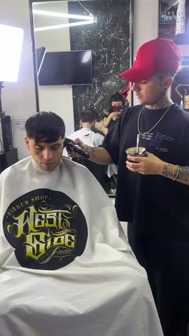 The video shows a man getting a haircut in a barber shop. The man receiving the haircut is wearing a white barber cape with a black and gold logo that reads "Barber Shop West Side Fade". He has short dark hair and tattoos on his face. He is sitting in a barber chair with his head slightly bowed.  The barber is wearing a red baseball cap, a black t-shirt, and black pants. He has tattoos on his arms and neck. He is using a hair clipper to cut the man's hair. The barber is also holding a metal cup with a straw in his left hand. There is a mirror behind the barber, reflecting the barber, the man getting a haircut, and another person in the background. In the reflection, the person in the background is wearing a mask and using a cell phone. There is a light panel to the left of the man getting his haircut. The barber shop appears clean and well-lit.