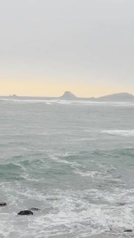 The video shows a choppy sea with waves crashing on a rocky shore. In the distance, several islands or large rocks can be seen jutting out of the water. The sky is overcast with a yellowish hue near the horizon, suggesting either sunrise or sunset. There are no people or man-made objects clearly visible in the frame. The overall atmosphere is moody and natural.