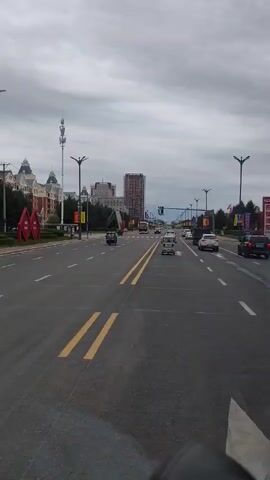 The video shows a view from a vehicle moving down a wide road. The road has multiple lanes and is marked with double yellow lines. Several cars and a blue three-wheeled vehicle are visible on the road. On the left side of the road, there are tall apartment buildings and decorative red diamond-shaped structures with yellow banners. Evergreen trees line the median. Streetlights are positioned along the road. The sky is overcast.