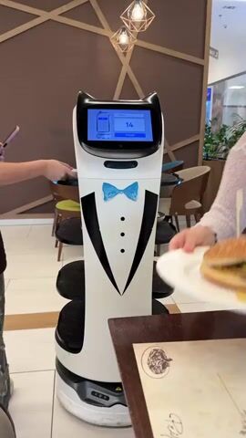 In this video, a robotic waiter, designed to resemble a tuxedo with a blue bowtie and cat ears, is shown in action within a restaurant. The robot has a screen displaying the number "14" and a "Finish" button. Several people are interacting with the robot. One person is placing a plate with a burger on it onto one of the robot's shelves. Another person is holding a phone and appears to be interacting with the robot's screen. A third person is placing a plate of french fries onto another shelf. The background shows a modern restaurant interior with tables, chairs, and some greenery. The lighting suggests it is daytime, likely indoors. The overall atmosphere appears casual and efficient, with people seemingly receiving their food orders from the robot. The video is shot from a close-up perspective, focusing on the robot and the immediate interactions around it. No specific time of day or weather is discernible, but the location appears to be an indoor dining establishment.