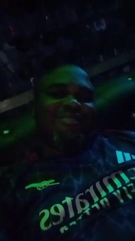 The video shows a dark-skinned man in a dark room, wearing an Arsenal football jersey with the Emirates logo. He's smiling and looking at the camera. The room is illuminated by colored flashing lights. There's a sound of music in the background. The camera appears to be hand-held and close to the man's face.